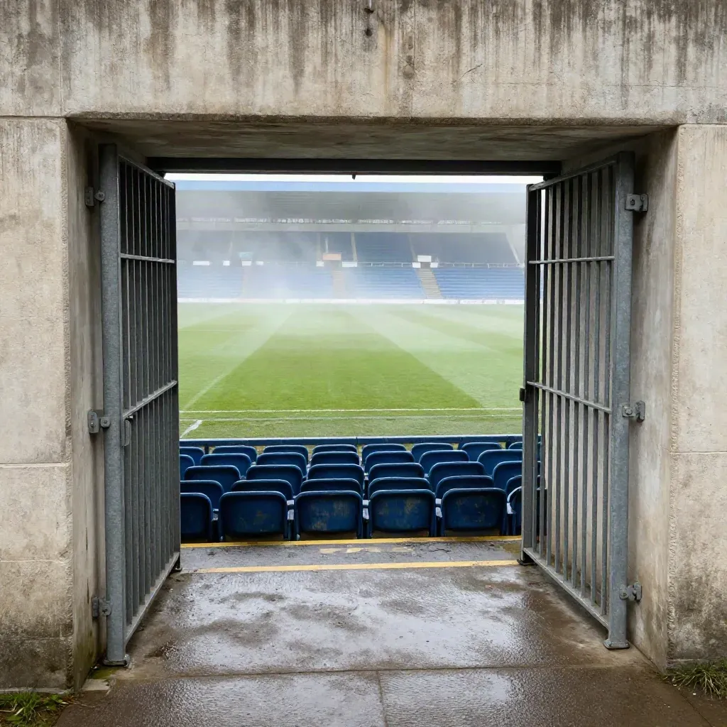 Tunnel dallo stadio verso il campo, cancelli metallici e gradinate blu nella luce del mattino.